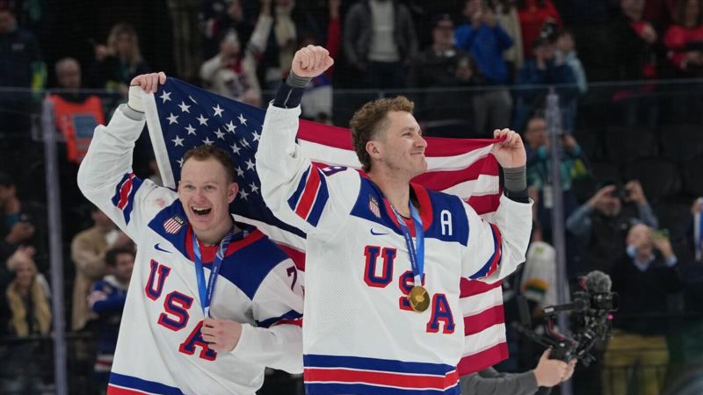 <i>NBC Olympics</i><br/>U.S. men's hockey players Brady and Matthew Tkachuk celebrate together on the ice after winning gold at the 2026 Milan Cortina Olympics with the American flag draped behind their backs and gold medals around their necks.