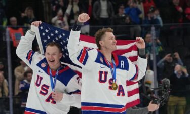 U.S. men's hockey players Brady and Matthew Tkachuk celebrate together on the ice after winning gold at the 2026 Milan Cortina Olympics with the American flag draped behind their backs and gold medals around their necks.