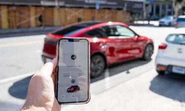 A smartphone displays the Tesla Robotaxi app while a Tesla Robotaxi Model Y self-driving vehicle arrives for pickup in San Francisco